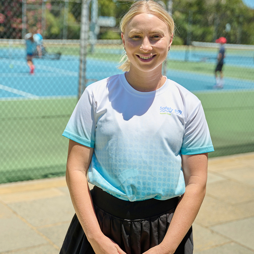 A woman with blonde hair tied back, wearing a white and blue Safety Bay t-shirt and a black skirt, stands smiling in front of a tennis court with players in the background.