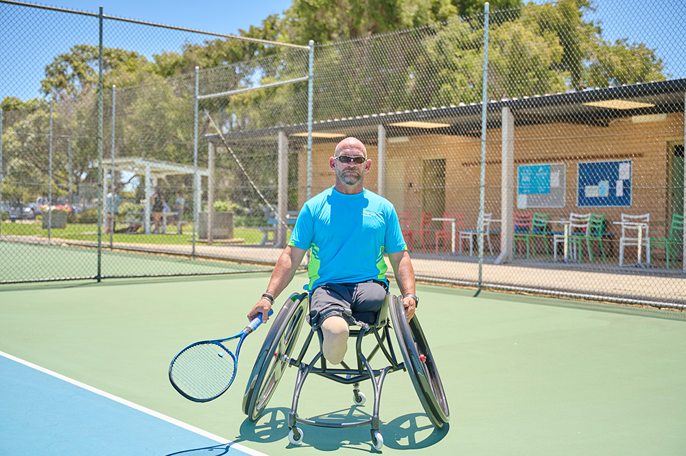 Man dressed in a light blue t-shirt and dark blue shorts and wearing sunglasses is holding a tennis racket in his hand. He is using a wheel chair.