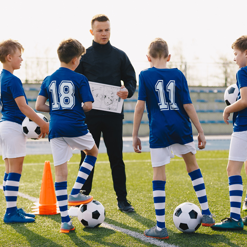 A coach holds a tactic board while giving instructions to four young boys in blue soccer uniforms, each holding a soccer ball, on a field with orange cones.