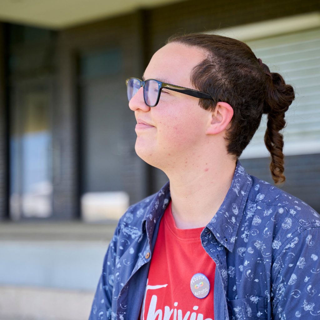 A person with glasses and a ponytail smiles while looking to the side. They wear a red shirt, a patterned blue overshirt, and a button badge, standing outdoors in front of a building with steps and large windows.
